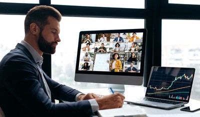 online-project-platform-for-host-companies A business owner working by his desk with an open laptop showing statistics during on online meeting with many virtual participants showing on the desktop screen.