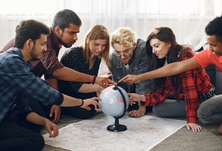 University students sitting on the floor with a world map and a globe all pointing at different locations on the globe.