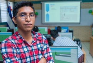 A science student is standing in a classroom with a science presentation projected on a white screen in the background.