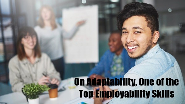A young, dark-haired, happy-looking intern by a meeting table wearing a collared shirt is looking behind him and towards the viewer during a team meeting while two colleagues are sitting by the meeting table and a lady on the blurred background is pointing to a flipchart.