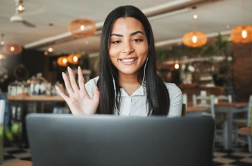 A young female graduate is sitting by her laptop in a cafe wearing headphones whilst taking part in a one-on-one online LinkedIn training session.